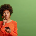Photo of pretty ethnic woman ponders on how to answer question, thinks deeply about something, uses modern mobile phone,...